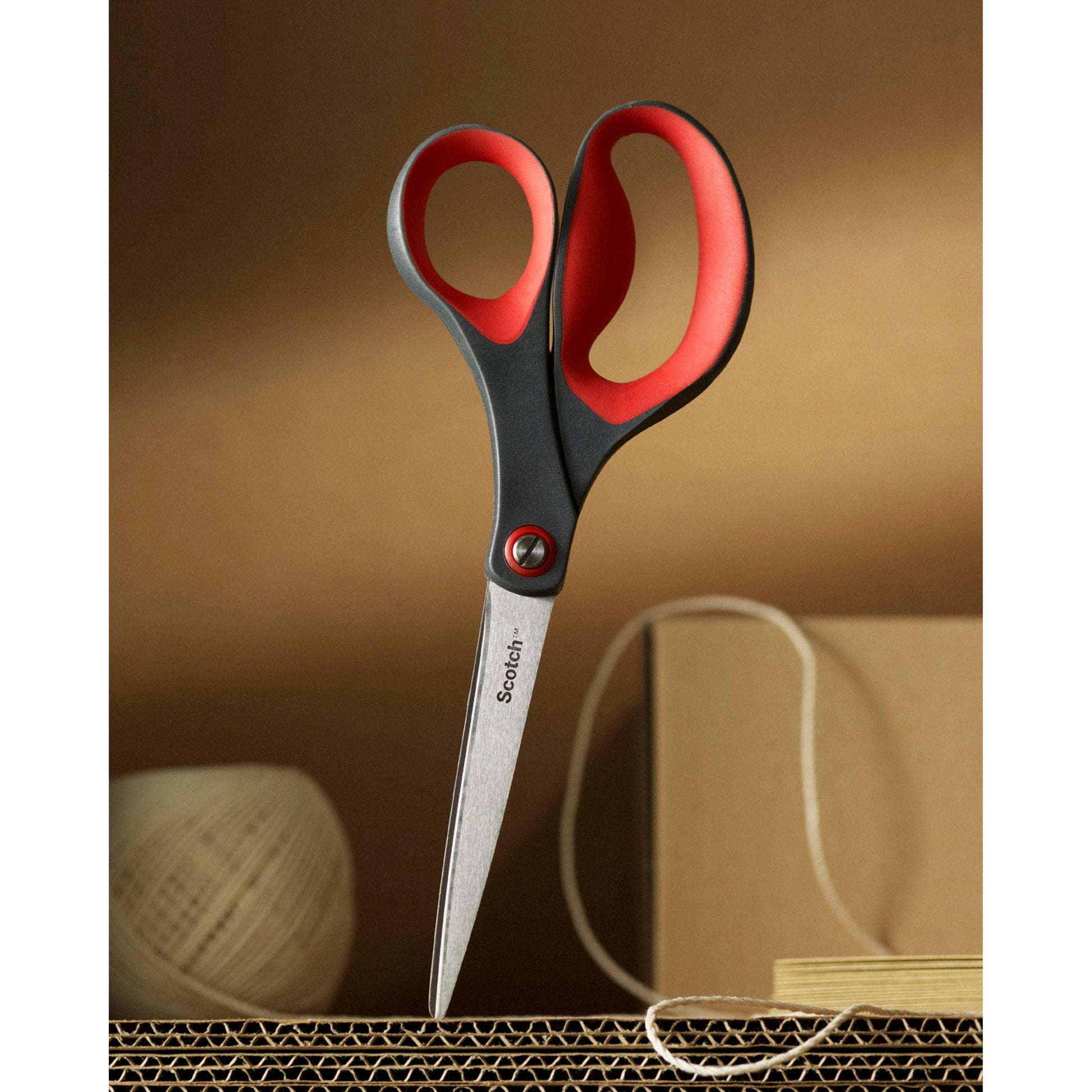 Ein Paar rote Scotch™ Präzisionsschere von 3M Deutschland GmbH ist aufrecht in Wellpappe geklebt, im Hintergrund sind beigefarbene Schnüre und ein Karton zu sehen.