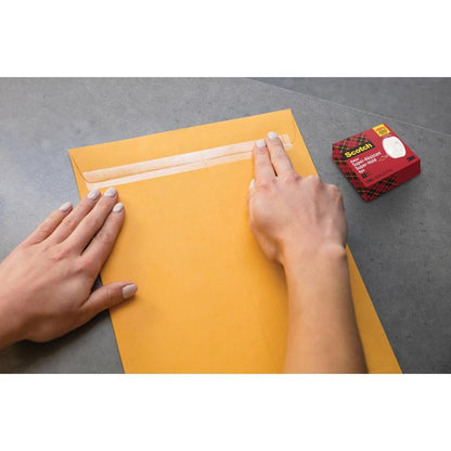 Henkilö sulkee suuren ruskean kirjekuoren Scotch® Super-Hold Secure Tape -teipillä, 1 rulla, 19 mm x 25,4 m 3M Deutschland GmbH:ltä harmaalla pinnalla. Teipin laatikko on lähellä ja takaa vahvan ja läpinäkyvän liimauksen turvalliseen postitukseen.