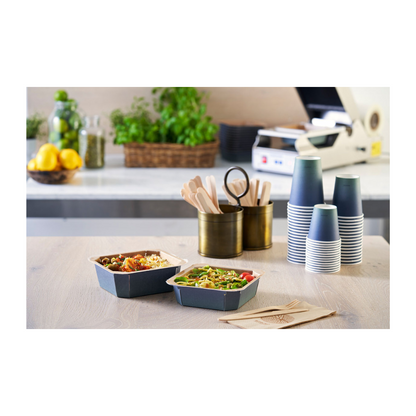 Pöydällä on kaksi take-away-kulhoa, joita reunustavat pinotut Duni Urban Kaffeebecher PAP/PLA -mukit Duni GmbH:ltä, puinen ruokailuvälinepidike ja lautasliinoja. Taustalla olevat sitruunat, lasit ja yrtit täydentävät tämän ruokavalmistusalueen kestävää ilmapiiriä.